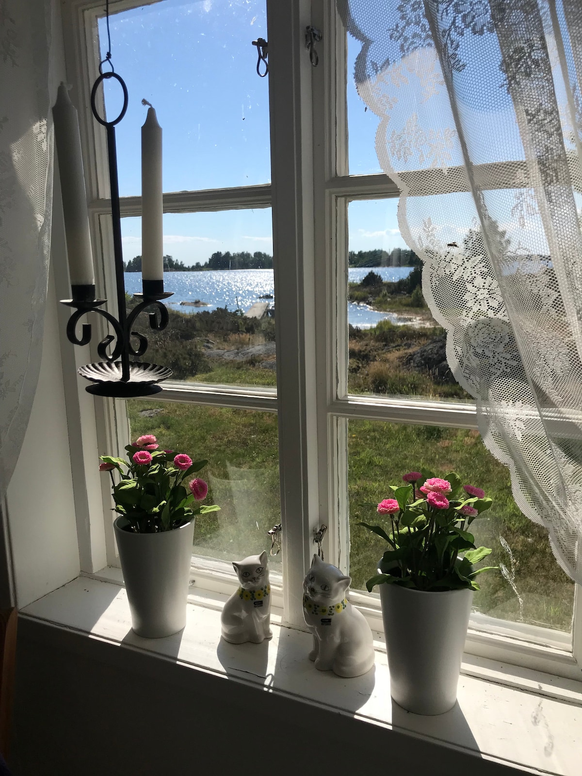 A window adorned with lace curtains offers a view of the natural landscape outside. Two white ceramic cats are positioned next to two potted plants with pink flowers on the windowsill, complemented by a candle holder hanging above.