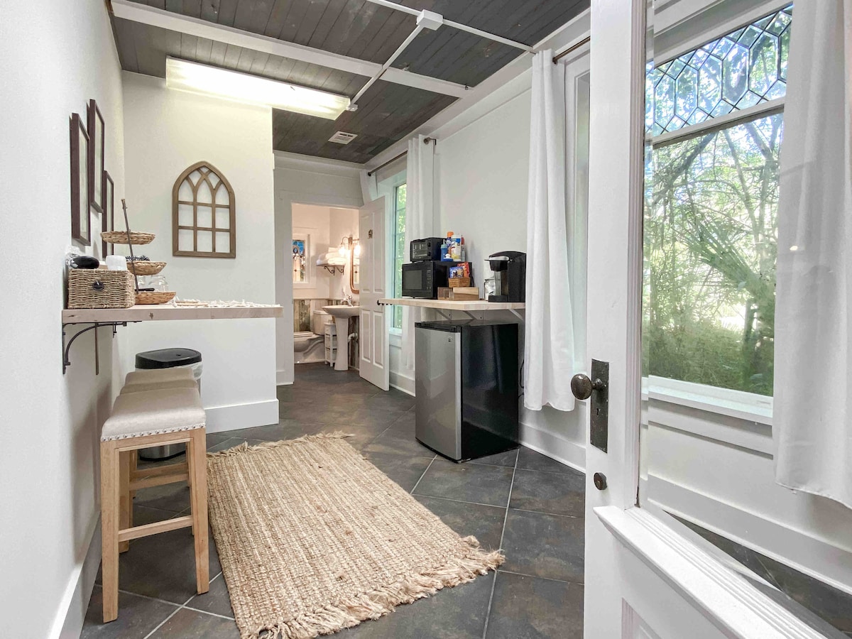 A kitchenette area is shown with a mini fridge and coffee maker. A small dining space is highlighted by a narrow table and two stools. Natural light is filtered through nearby windows featuring decorative glass panes, enhancing the space's connection to the outdoor environment.