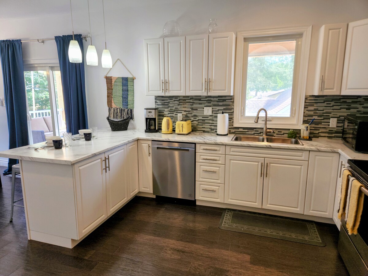 A well-organized kitchen is equipped with modern appliances, including a stainless steel dishwasher and a yellow kettle. Light wood cabinetry contrasts with the dark countertops, while a window provides natural light and a view of the outdoor area. Dining essentials are arranged on the countertop.