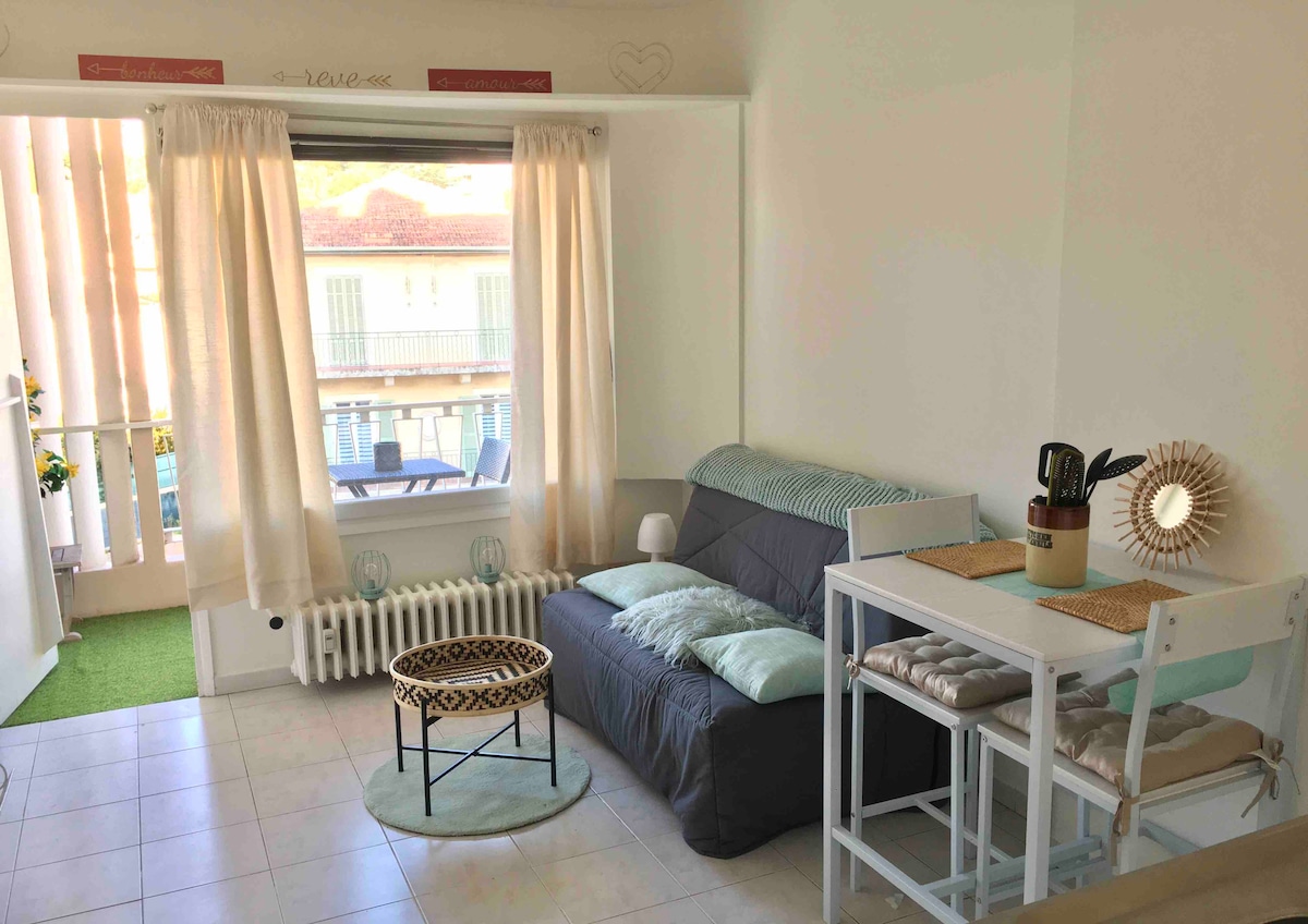 A cozy living area features a comfortable sofa bed adorned with soft cushions. A small circular table is situated in front, accompanied by two bar stools. Natural light fills the space through large windows draped with light curtains, offering a view of a terrace.