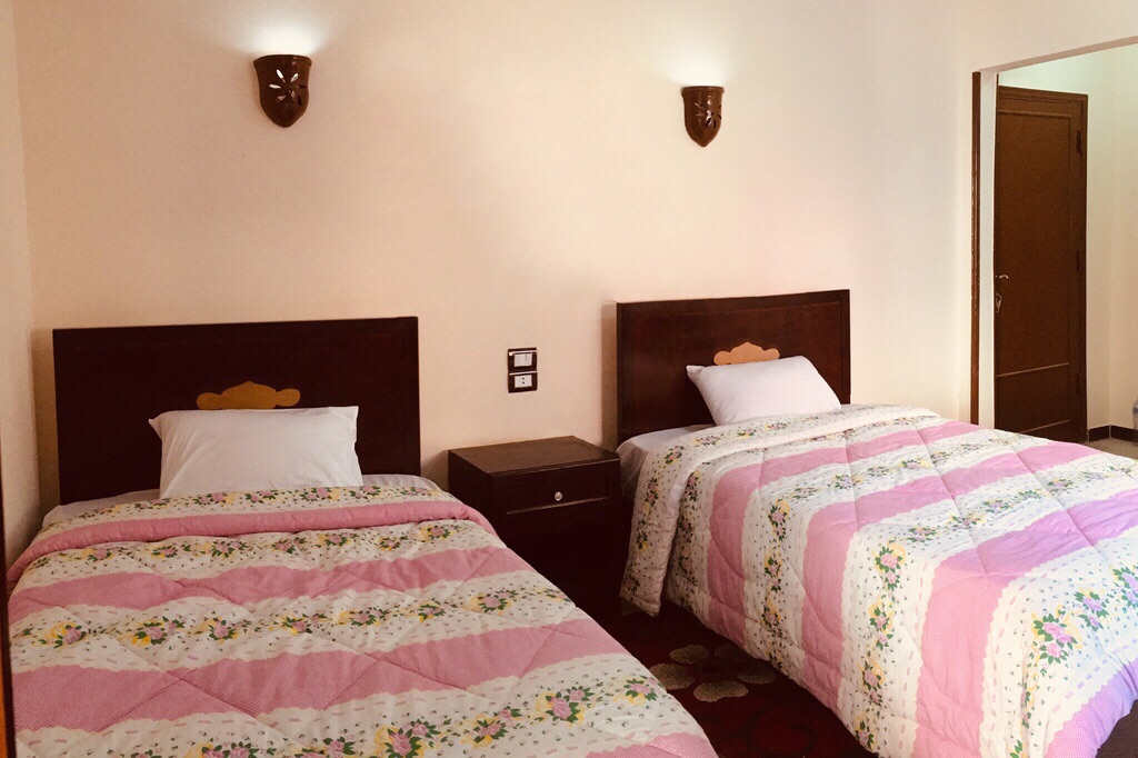 A spacious twin room is shown, featuring two beds with floral patterned quilts. A nightstand is positioned between the beds, and minimalist wall sconces provide soft lighting. An open doorway is visible in the background, leading to additional areas of the guesthouse.