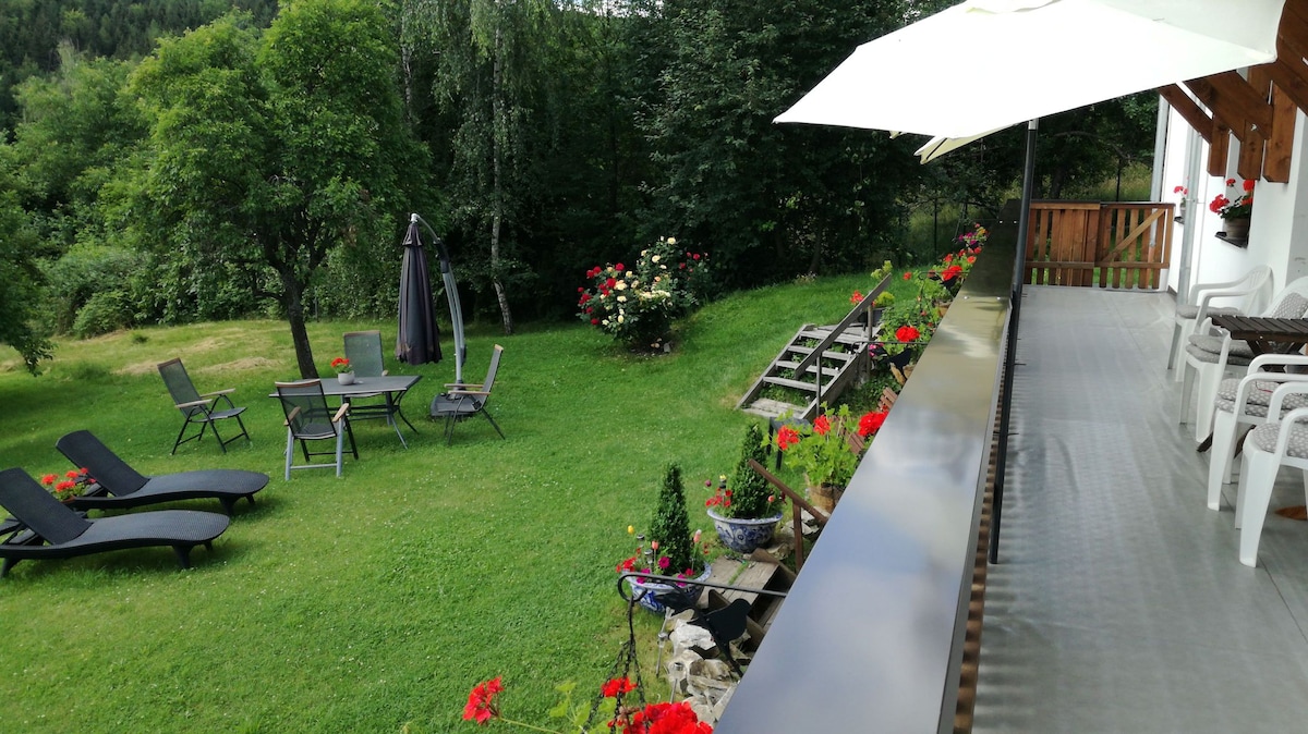 A spacious balcony extends along the house, offering a view of a lush garden. The area below features a seating set with lounge chairs and an umbrella, surrounded by manicured grass and blooming flowers. A pathway leads to additional seating areas beneath trees.