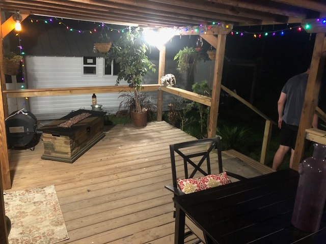A spacious back porch is adorned with colorful string lights, providing a warm ambiance. A wooden table and chairs are set on the wooden floor, complemented by a decorative rug. A grill stands nearby, with plants enhancing the welcoming space, and a gentle night view of the surroundings.