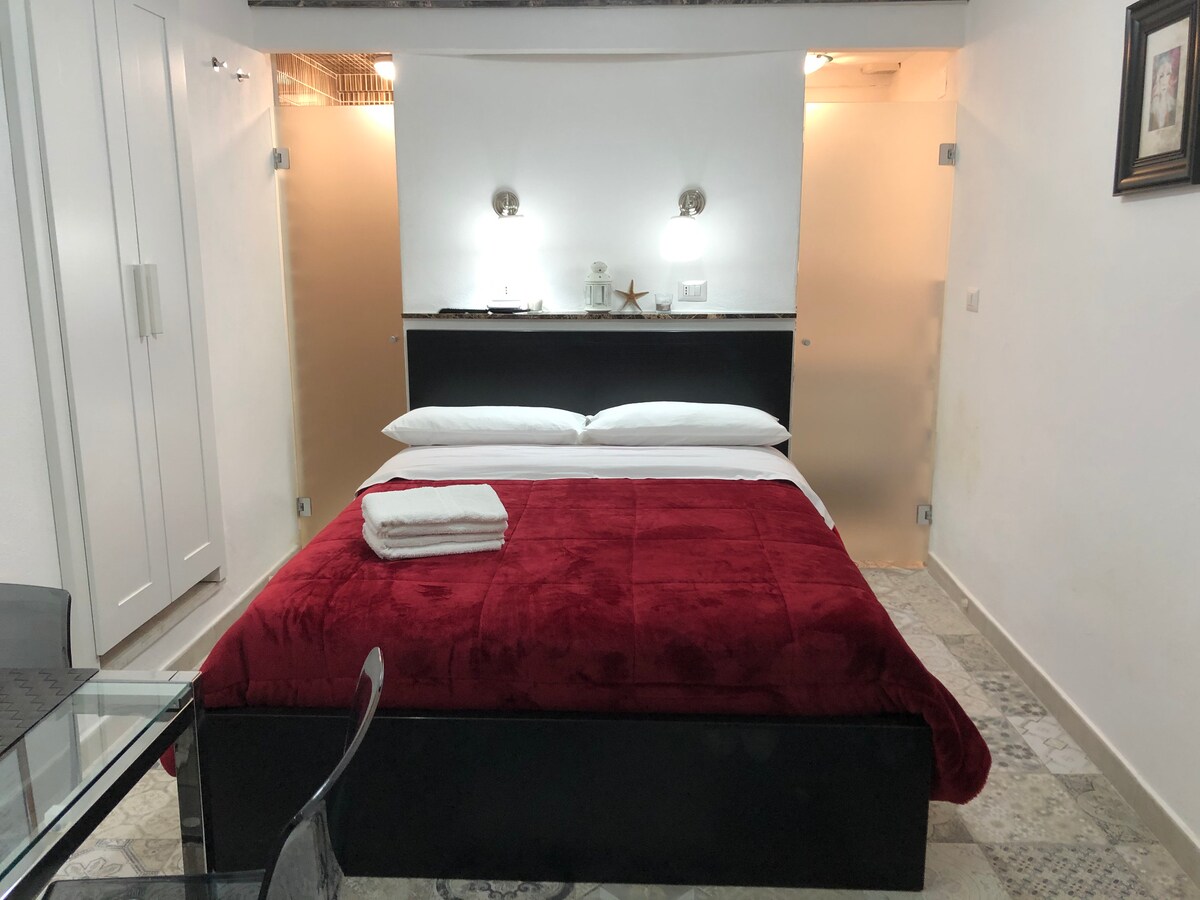 A sleeping area features a large bed with a soft red blanket and neatly arranged white linens. Soft lighting is provided by wall sconces above the bed. A glass table and chairs are positioned nearby, while light-colored cabinets are visible in the background.