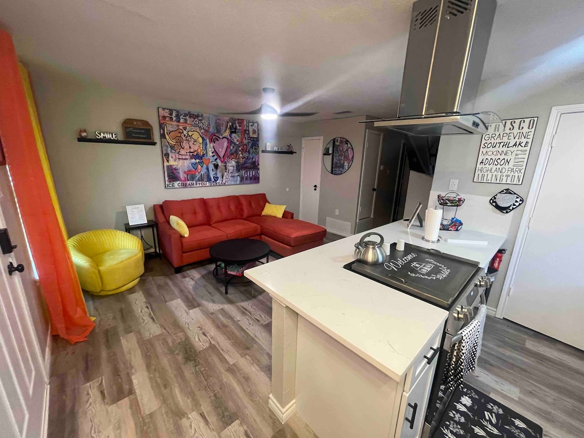 An open living space features a red sectional sofa, a round black coffee table, and a bright yellow bean bag chair. A well-equipped kitchen is observed in the background, highlighted by a stainless steel range hood and modern countertops. Colorful wall art adds a creative touch.