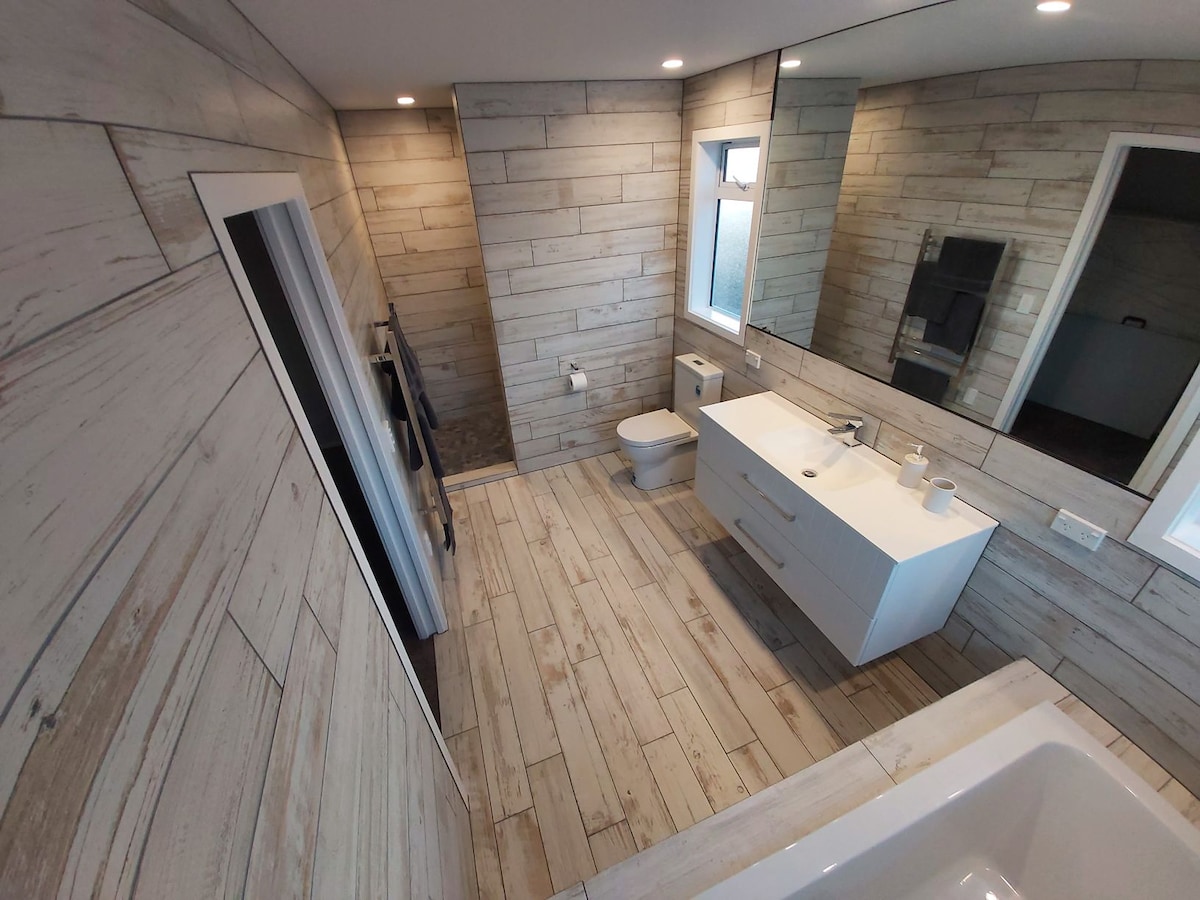 The spacious bathroom features light-colored wood paneling, a modern vanity with dual sinks, and a large mirror reflecting natural light. A sleek toilet is positioned to one side, while a separate shower area is visible. A bathtub is situated in the opposite corner, adding functionality.