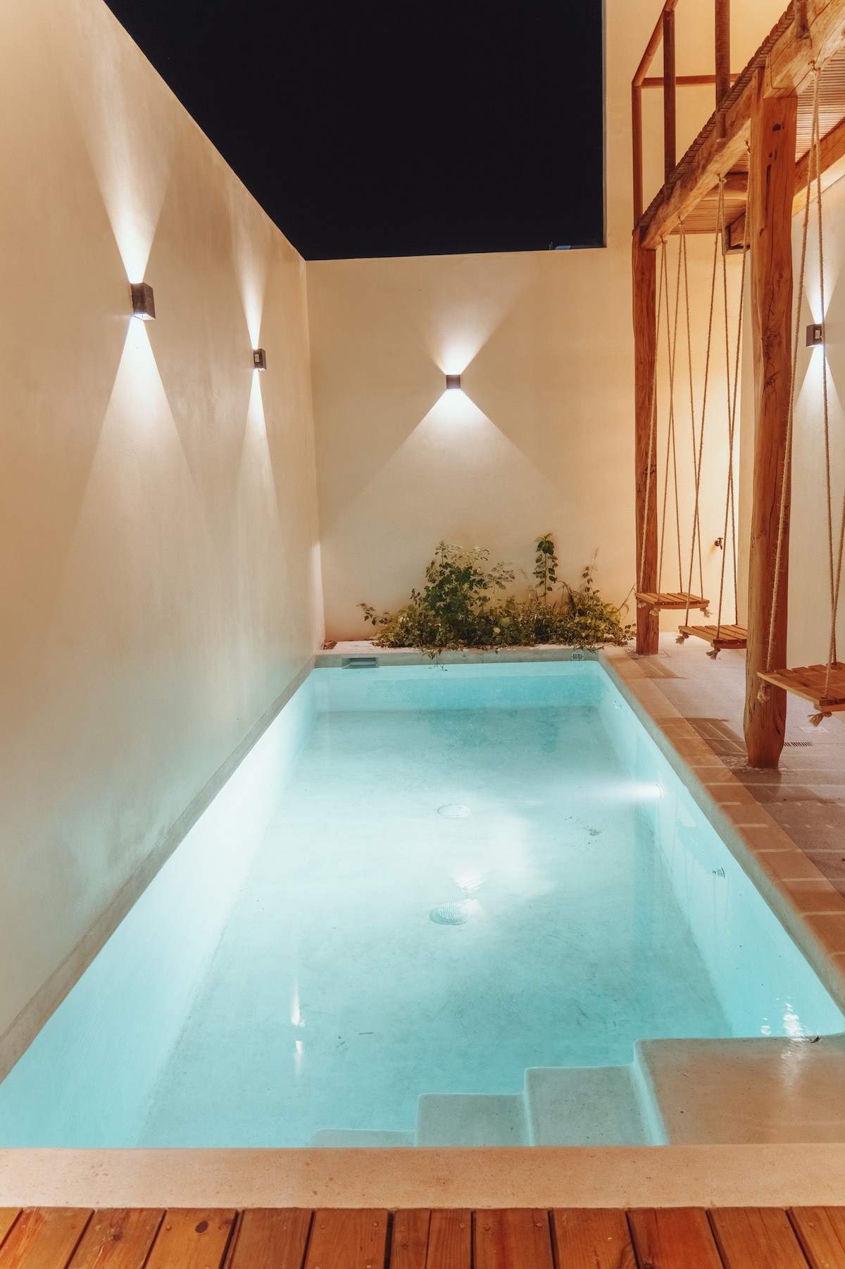 A small swimming pool with clear blue water is surrounded by smooth, light-colored walls. Soft lighting is positioned along the walls, creating a serene atmosphere at night. Wooden decking offers an inviting area for relaxation alongside the pool's edge.