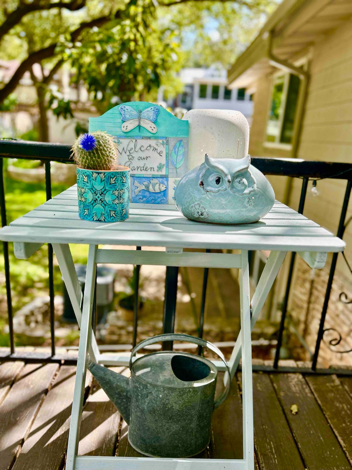 A small, white table is positioned on the deck, decorated with a variety of items. A blue-painted owl figurine and a turquoise planter with a cactus sit alongside a decorative sign welcoming guests. A silver watering can rests below, adding to the garden theme.