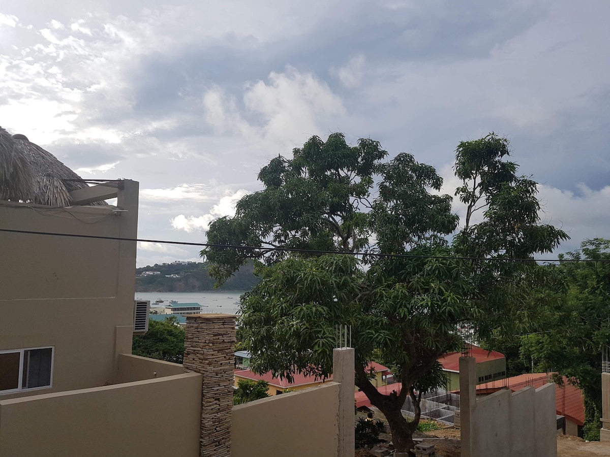 A view from the balcony presents a large green tree with lush foliage beside a sandy area. The ocean glistens in the background, and rooftops of nearby buildings are visible, contributing to a serene coastal atmosphere.