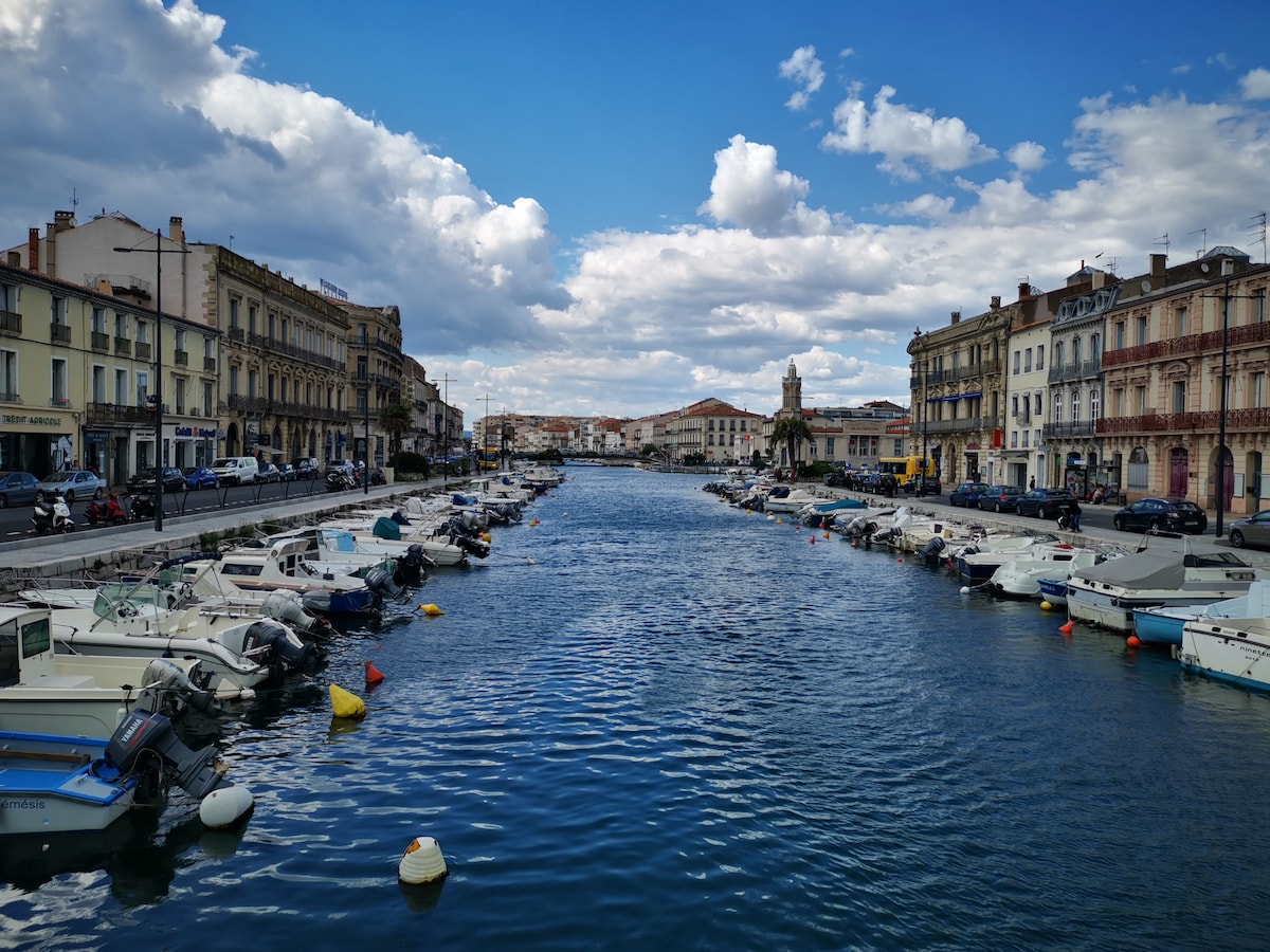 A serene view of a canal is featured, lined with small boats. The clear blue water reflects the sky dotted with clouds. Historic buildings flank both sides of the waterway, showcasing a blend of architectural styles that characterize the area.