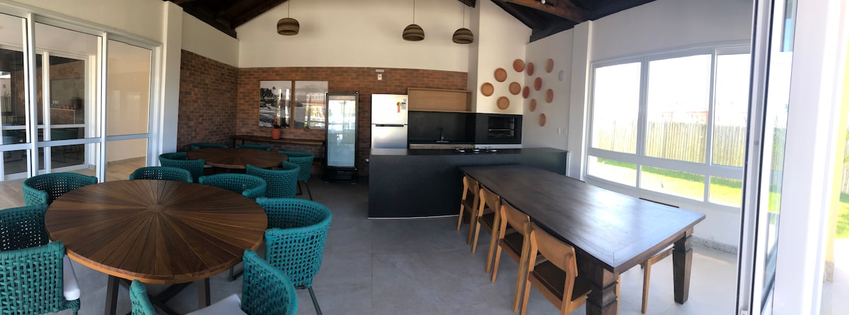 A spacious gourmet area is showcased with a large wooden dining table surrounded by comfortable seating. The kitchen features sleek appliances and a modern design. Large windows allow light to fill the space, creating an inviting atmosphere with views of the outdoor area.
