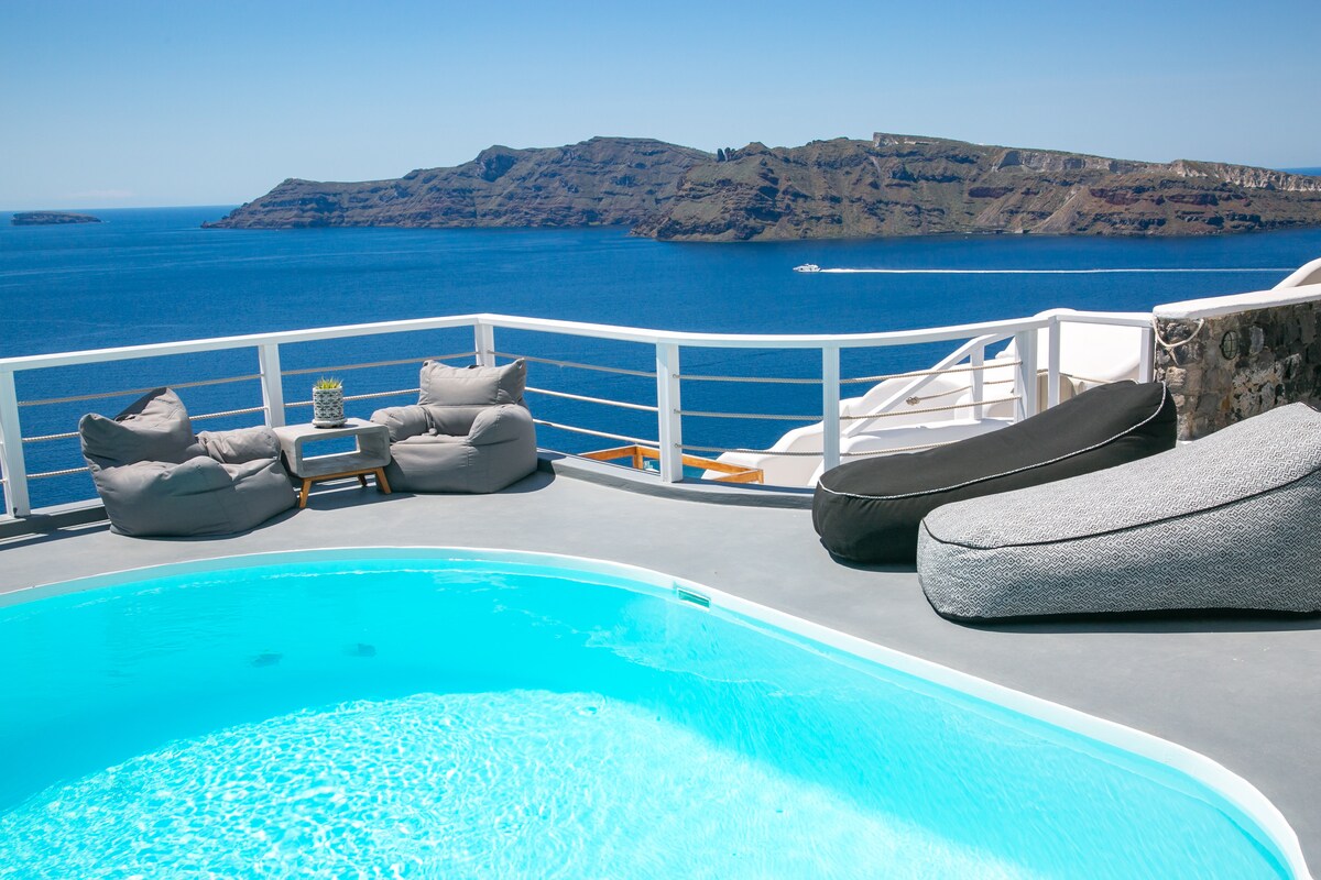 A private terrace features a refreshing plunge pool surrounded by cozy bean bags. The expansive views reveal the serene Aegean Sea and volcanic cliffs, providing a tranquil backdrop for relaxation. Bright blue waters sparkle under the clear sky, enhancing the inviting outdoor space.
