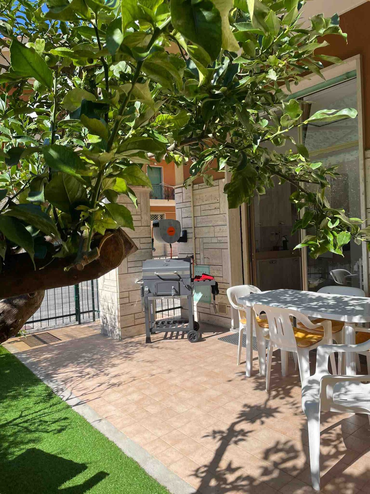 A private outdoor area features a shaded seating arrangement with a table and chairs, complemented by a barbecue grill. A lush green tree adds natural shade, creating a cozy spot for dining or relaxation. The area is enclosed for added privacy.
