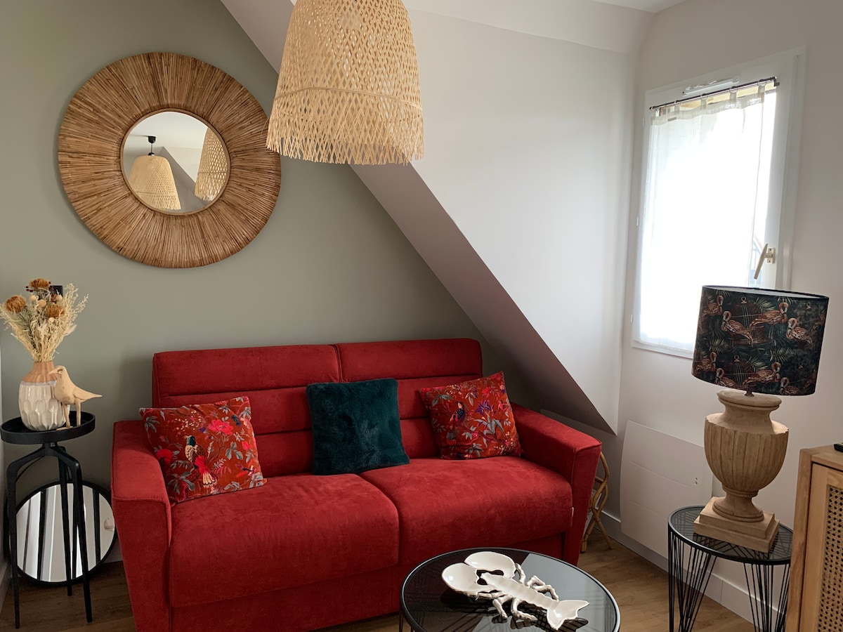 A cozy living area is shown with a bright red sofa adorned with decorative pillows. A round mirror reflects natural light, and a rattan lampshade adds texture. A glass coffee table is placed centrally, accompanied by a small side table and a unique lamp.