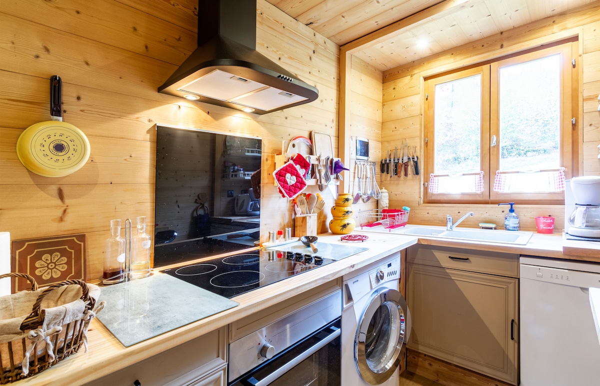A well-equipped kitchen features wooden walls and a window allowing natural light. An induction cooktop and a modern oven are complemented by a washing machine. Various utensils and kitchen tools are organized on a wall rack, contributing to a functional cooking space.