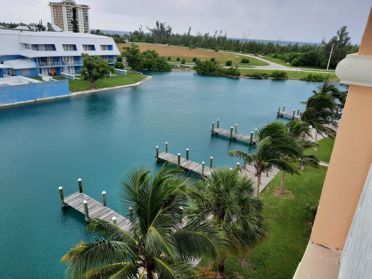A serene waterfront view is displayed, featuring calm turquoise waters lined with wooden docks. Lush green palm trees sway gently along the shoreline, complemented by manicured grass. The background reveals buildings and greenery, enhancing the tranquil atmosphere of the location.