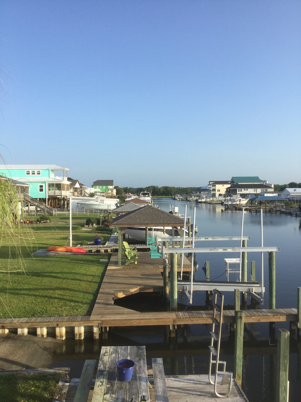 The image features a canal lined with grassy areas and wooden docks. Colorful houses are visible on the opposite bank. Two kayaks can be seen on the dock, while a gazebo offers shaded seating. The water reflects the clear blue sky.