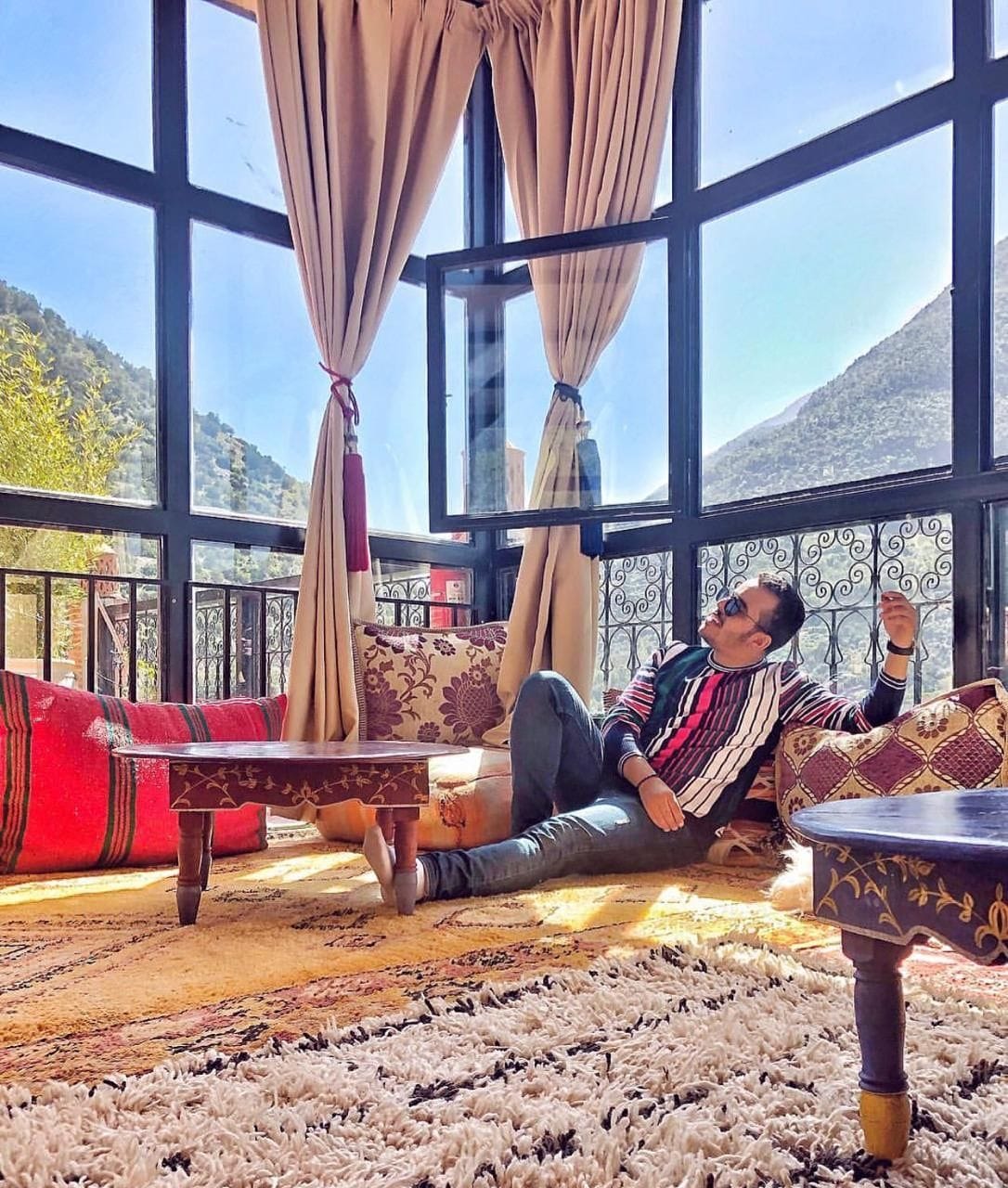 A sunlit lounge area features large windows offering a panoramic view of the mountainous landscape. Richly patterned cushions are arranged on a plush rug, alongside small wooden tables, creating a cozy seating space. Drapes frame the windows, enhancing the bright ambiance.