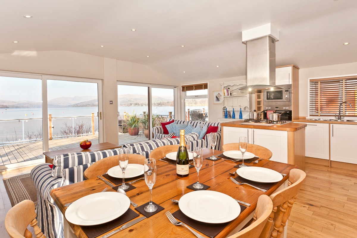An open-plan dining area features a wooden table set for four, with plates and glasses in place. The kitchen area, equipped with modern appliances, is adjacent to a comfortable seating area with striped sofas. Large windows provide a view of the lake and mountains.