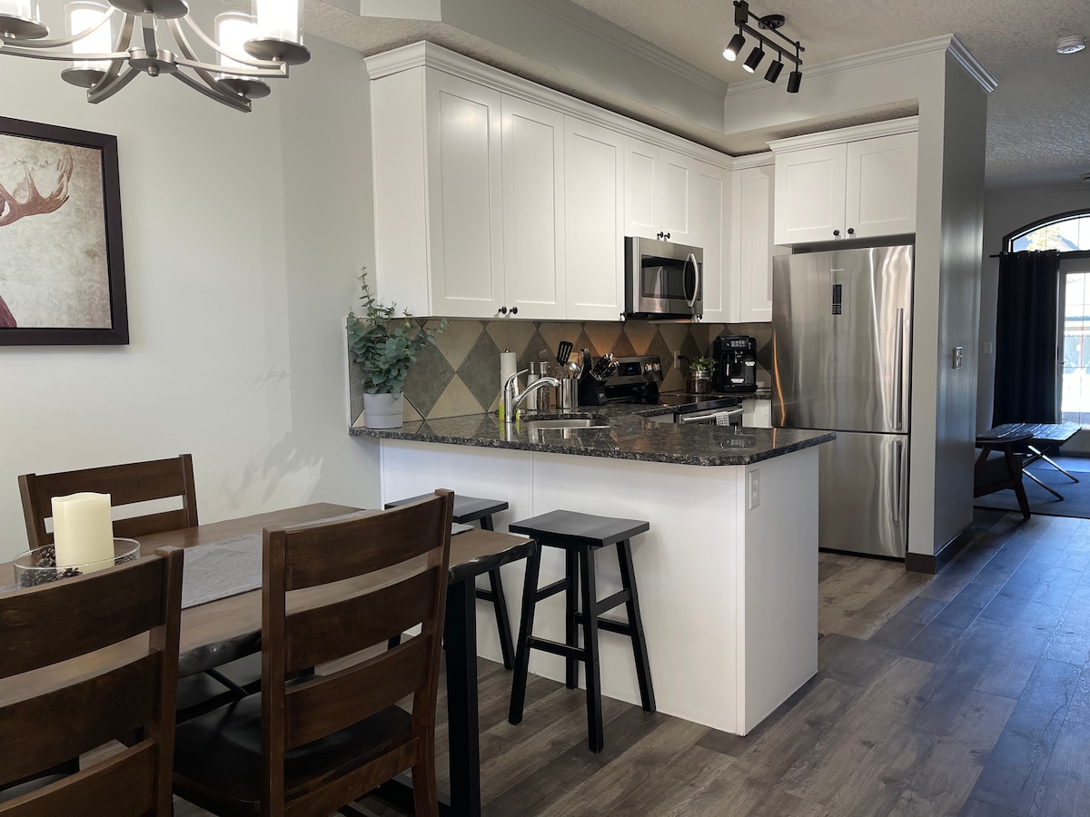 A fully equipped kitchen is showcased with modern appliances, including a refrigerator and microwave. A dining table with dark wood chairs is positioned nearby. Soft lighting enhances the welcoming atmosphere, and bar stools offer additional seating at the kitchen counter.