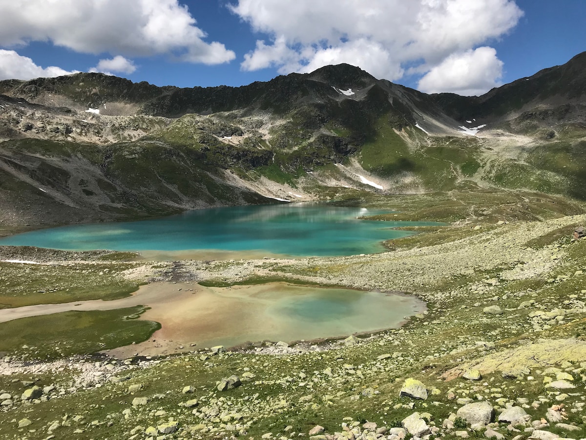 A glistening alpine lake nestled among rugged mountains is displayed. The water reflects varying shades of turquoise, surrounded by grassy banks and scattered rocks. Distant peaks are capped with snow, contrasting against a partly cloudy sky.