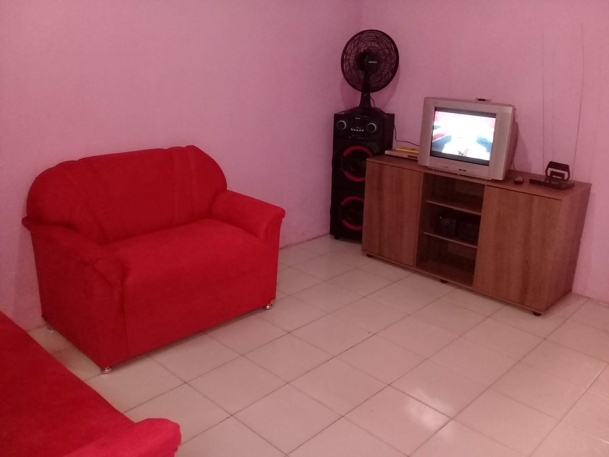 A living area features a red sofa and a matching loveseat set against pink walls. A television is positioned on a wooden media cabinet, alongside a speaker system, creating an inviting space for relaxation.