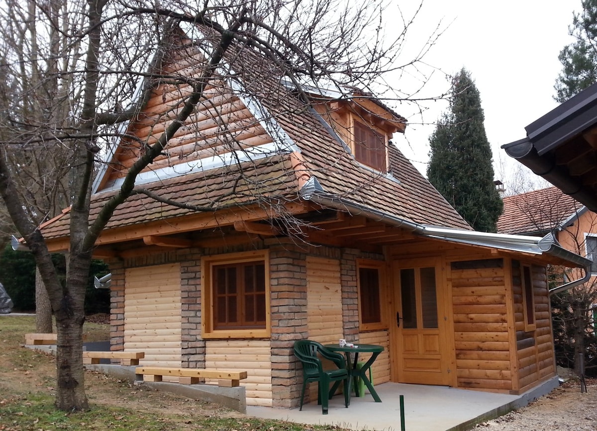 The exterior of a charming wooden chalet is featured, showcasing a combination of log and stone finishes. A sloped roof and multiple windows enhance its rustic appeal. A small patio area is equipped with plastic chairs, surrounded by trees and green grass.