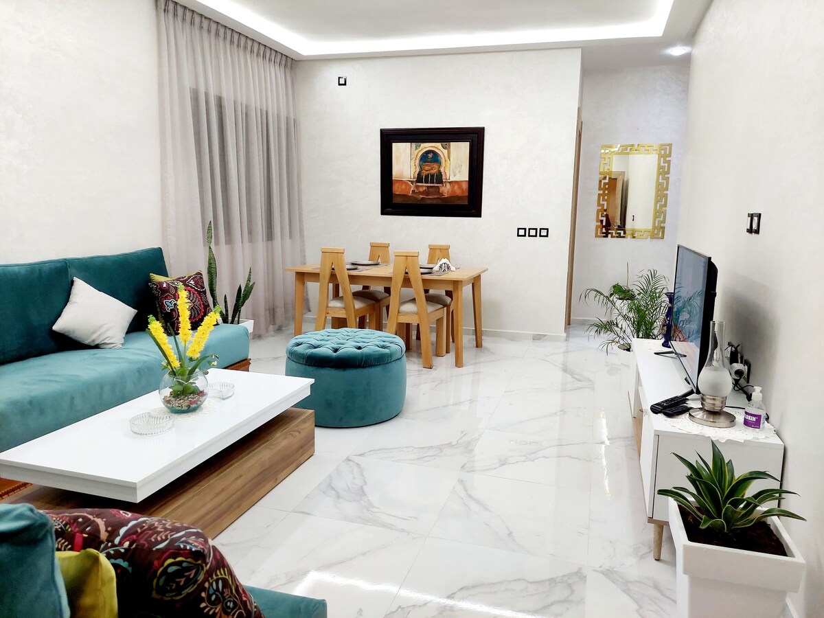 A modern living area features a comfortable teal sofa and matching ottoman. A white coffee table rests on polished marble flooring, while a dining table with wooden chairs and decorative plants accents the space. Natural light filters through sheer curtains, illuminating the design.