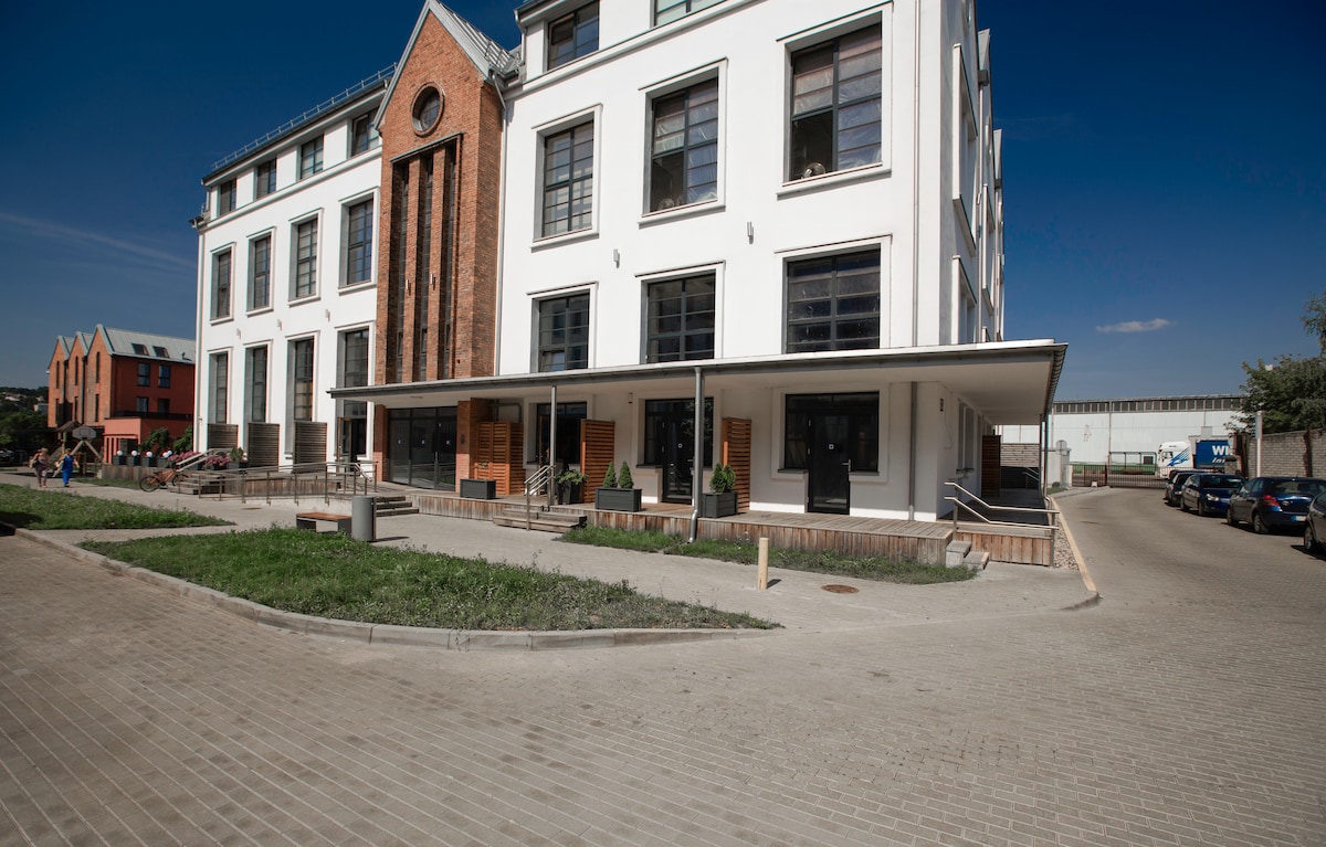 The exterior of a modern apartment building exhibits a blend of classic and contemporary design. Large windows align symmetrically with the flat façade, while a distinctive brick feature stands at the center. A landscaped area is visible in the foreground, with paved pathways leading to the entrance.