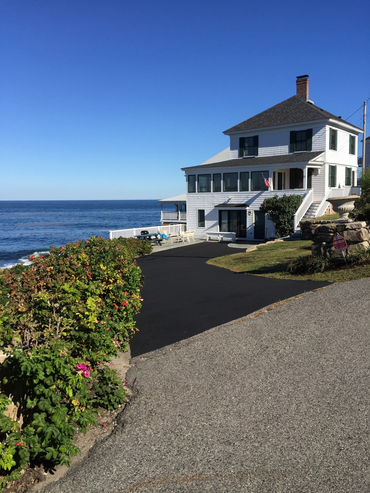 The charming cottage is positioned along the rocky coastline, overlooking the Atlantic Ocean. A well-maintained path leads to the entrance, with colorful greenery bordering the driveway. The clear blue sky completes the coastal scene, providing an inviting view of the structure and surrounding landscape.