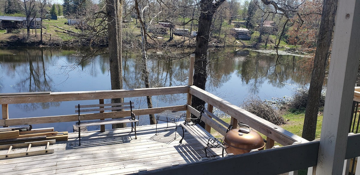 An outdoor deck overlooks the calm waters of a lake, framed by trees. A small table and chairs are positioned on the deck, providing a space to enjoy the view. The reflections on the water and the surrounding greenery create a serene setting.