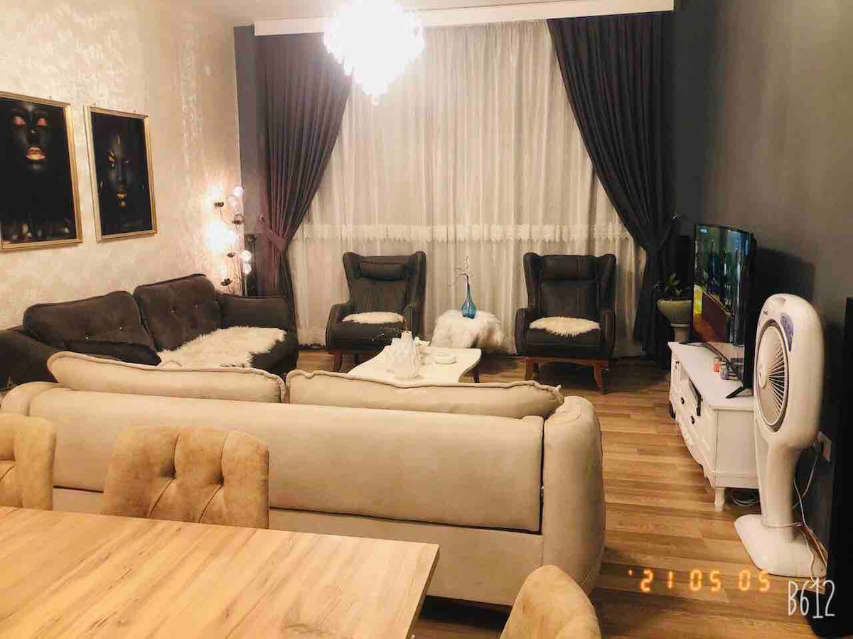 A well-furnished living area is showcased, featuring a comfortable sectional sofa and two armchairs. A sleek coffee table is positioned centrally. A television is mounted on a white console, while decorative lighting accents the space, complemented by floor-to-ceiling curtains framing the large window.