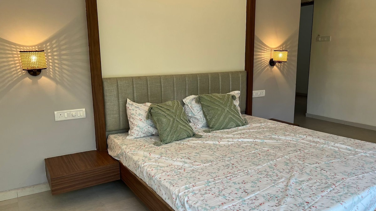 A spacious bed with a light floral-patterned covering is positioned against a wall. Two decorative pillows add subtle design touches. Soft wall sconces illuminate the area, creating a calm ambiance in a room with neutral tones.
