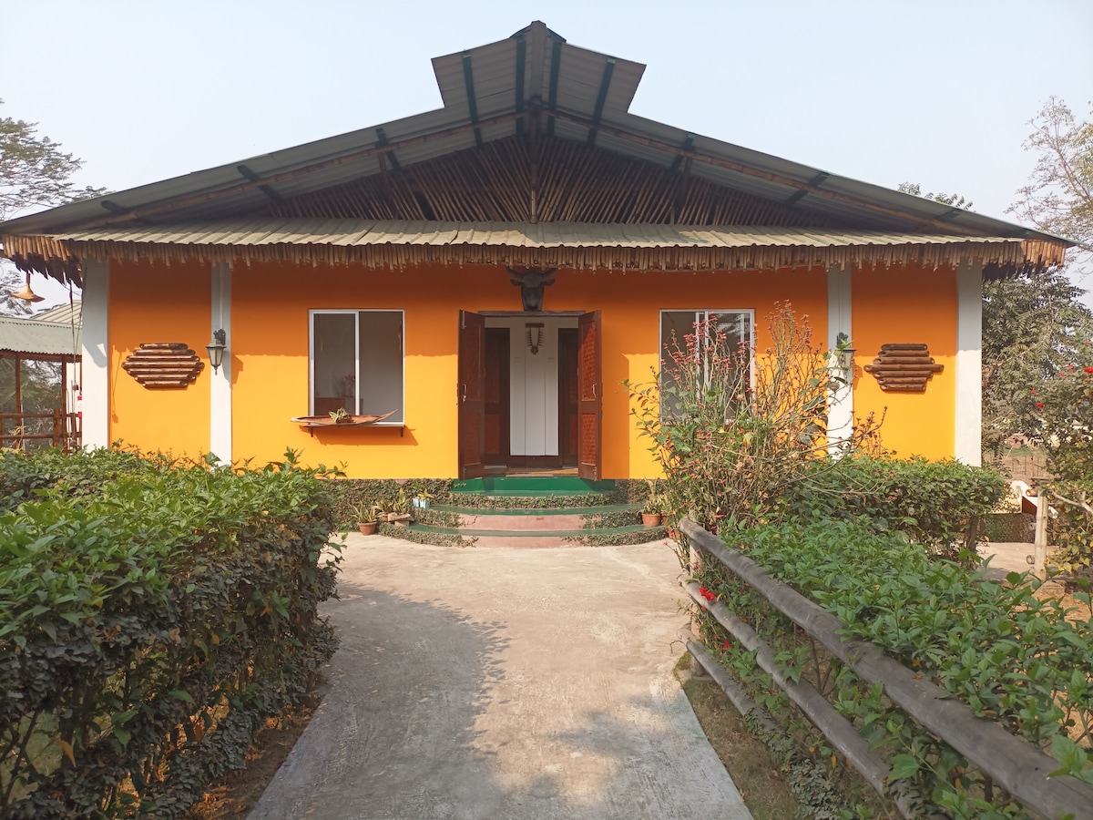 The entrance of a vibrant orange-stucco building is framed by a well-maintained garden. A concrete walkway leads to the front door, flanked by two windows. The roof is gently sloped with decorative elements, providing a welcoming appearance.
