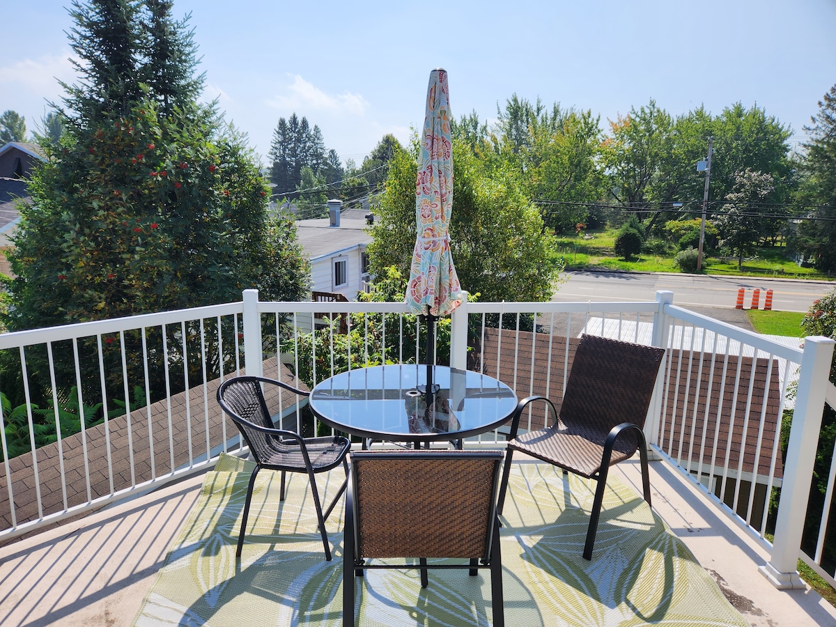 An outdoor patio area is equipped with a circular glass table surrounded by four wicker chairs. A colorful umbrella provides shade, and a patterned rug adds comfort underfoot. Lush greenery and trees frame the view, contributing to a serene atmosphere.