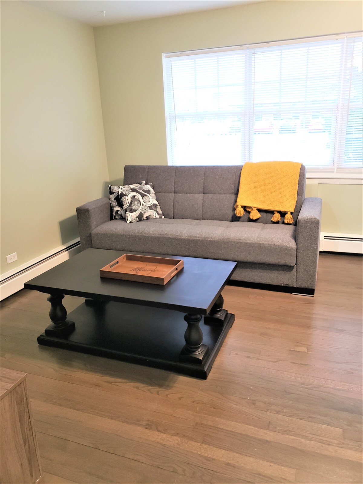 A comfortable seating area features a gray sofa adorned with a yellow throw blanket and decorative pillows. A dark wood coffee table with a wooden tray occupies the center, while natural light filters in through a window adorned with horizontal blinds.