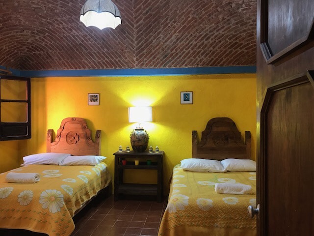 A brightly colored room is featured with two spacious beds, each adorned with yellow bedspreads. A wooden nightstand sits between them, holding a decorative lamp and a ceramic vase. The textured brick ceiling and warm yellow walls enhance the inviting ambiance.