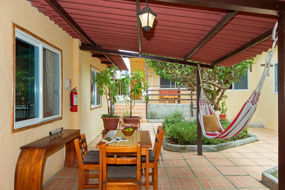 An inviting outdoor area features a wooden dining table with two benches under a protective overhang. A comfortable hammock is hung nearby, and lush greenery adds a natural touch to the surroundings. The space is well-lit by a hanging lantern.