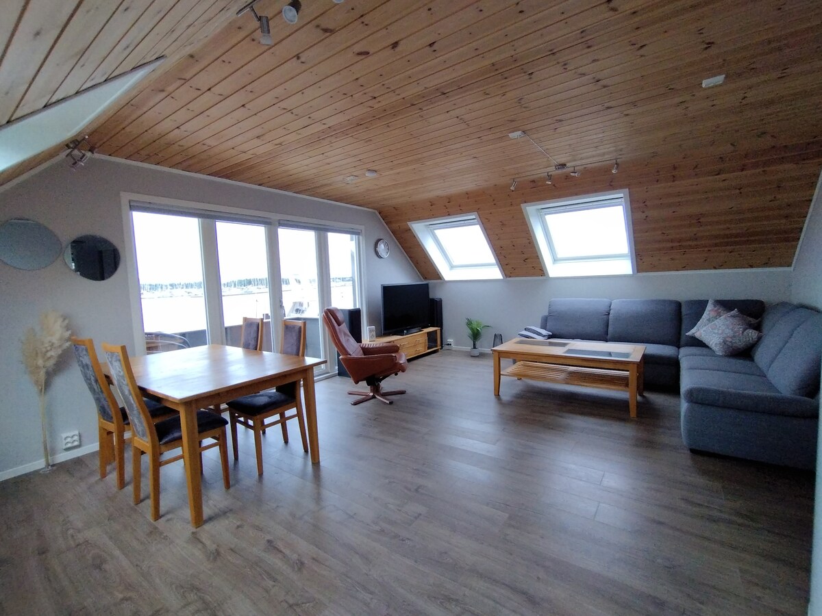 A spacious living area features wood-paneled ceilings and large windows allowing natural light to fill the space. A comfortable sofa and a wooden coffee table are arranged around a television. A dining table accommodates four people, enhancing the communal atmosphere.