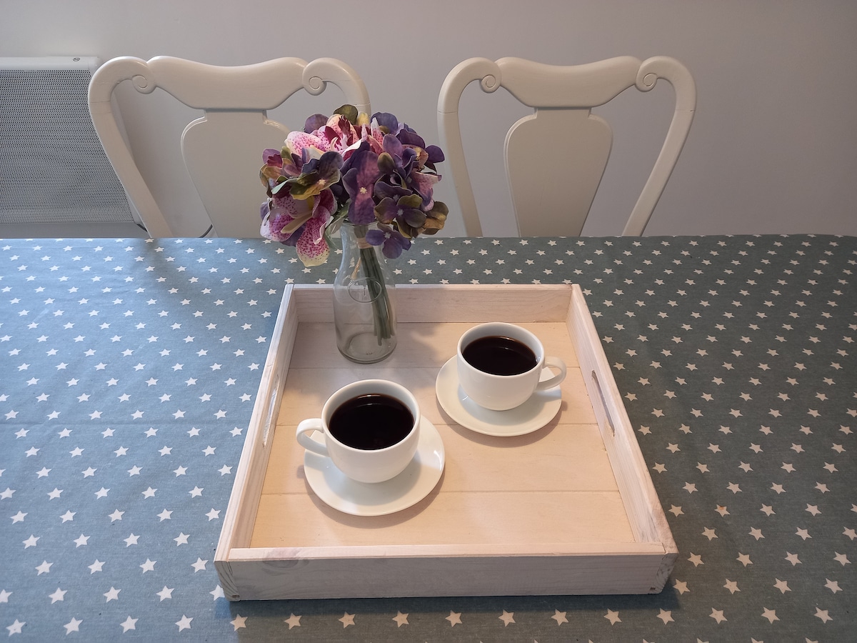 A wooden tray with two cups of coffee is placed on a table covered with a star-patterned cloth. A small glass vase holds a bouquet of purple flowers, adding a touch of color to the setting. Two white chairs are positioned at the table.
