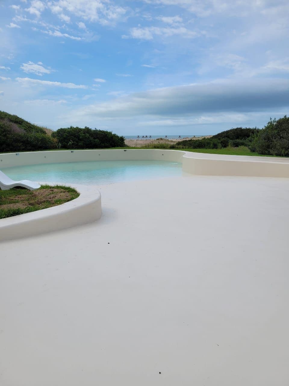 A tranquil pool area is visible, surrounded by neatly maintained grass and shrubbery. The smooth white surface of the deck contrasts with the serene water. In the background, a view of the sea and distant greenery can be seen under a cloudy sky.