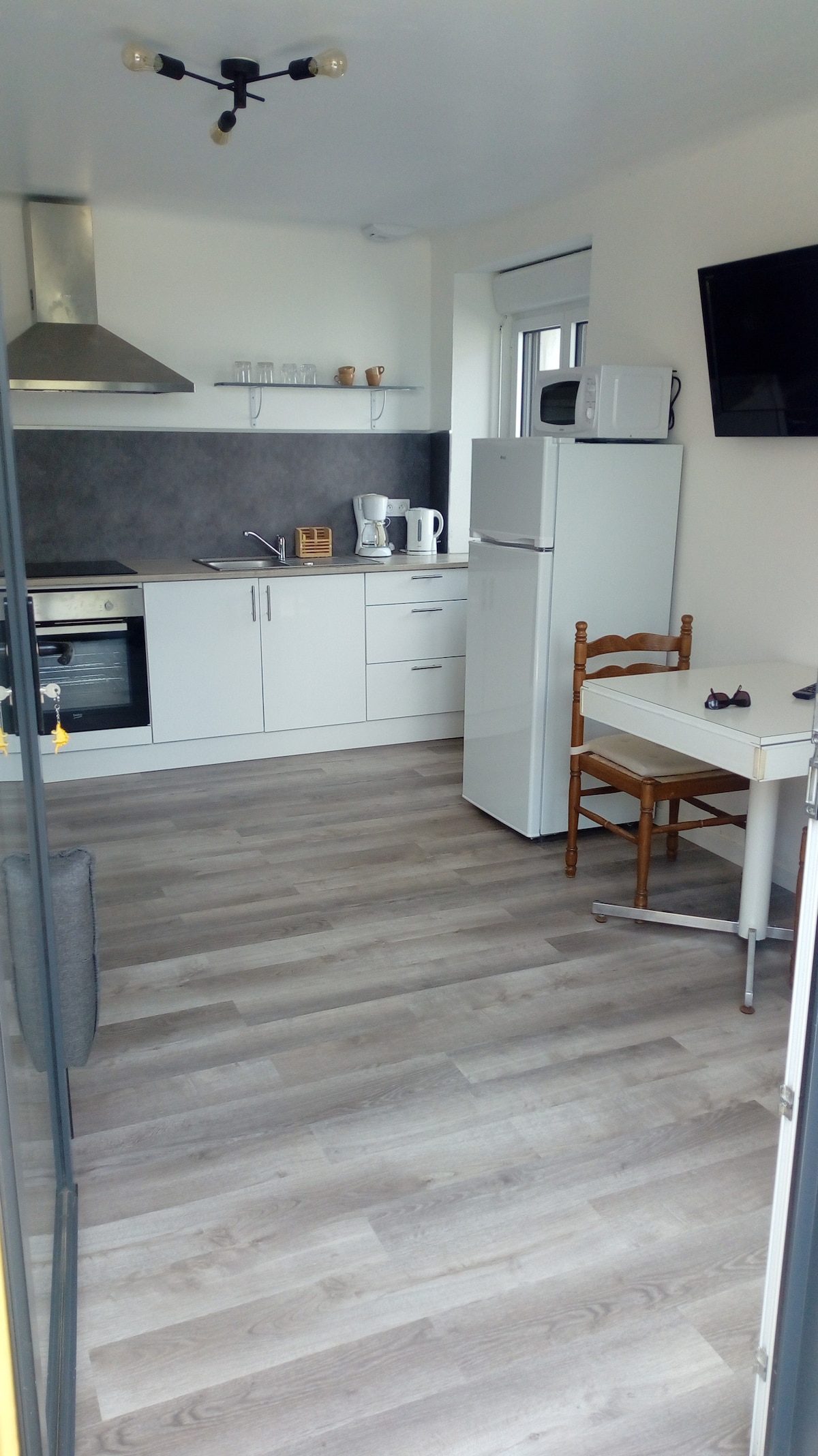 The kitchen area features modern appliances, including an oven, stovetop, refrigerator, and microwave. A small dining table with wooden chairs and a compact living space with a couch are visible. Natural light floods in through the windows, enhancing the light-colored flooring.