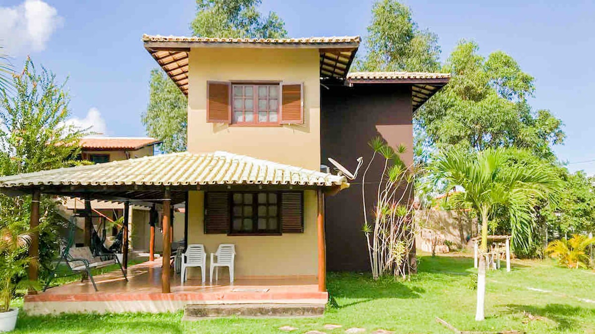 A two-story house with a tile roof and wooden shutters is set within a grassy area. A covered porch features two chairs for seating, and surrounding greenery enhances the rural ambiance. The exterior showcases a blend of earthy tones and natural elements.