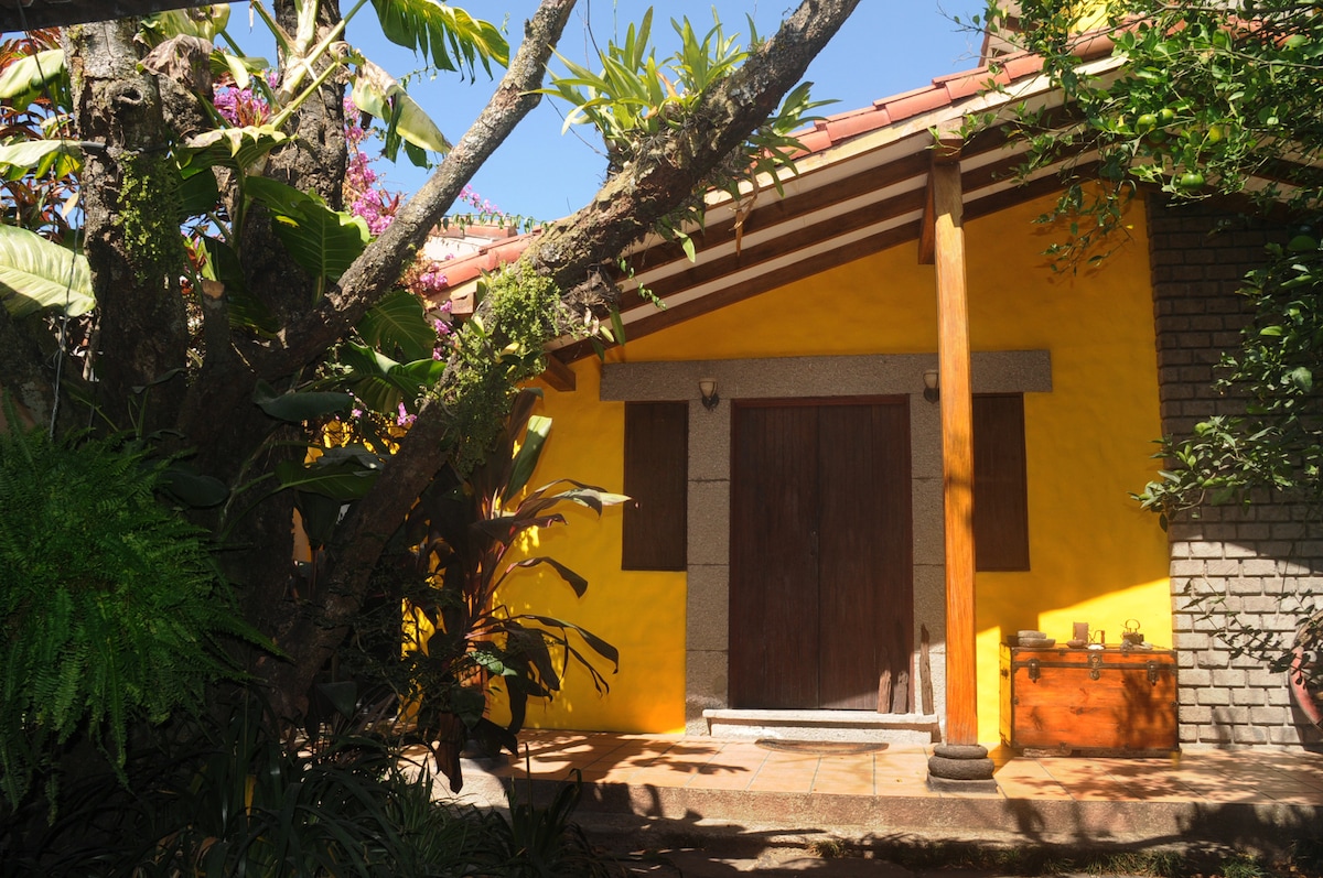 A bright yellow exterior features wooden doors and stone accents, surrounded by lush greenery and vibrant plants. The inviting entrance is framed by natural elements, creating a serene atmosphere. Sunlight filters through the leaves, highlighting the textures and colors of the space.