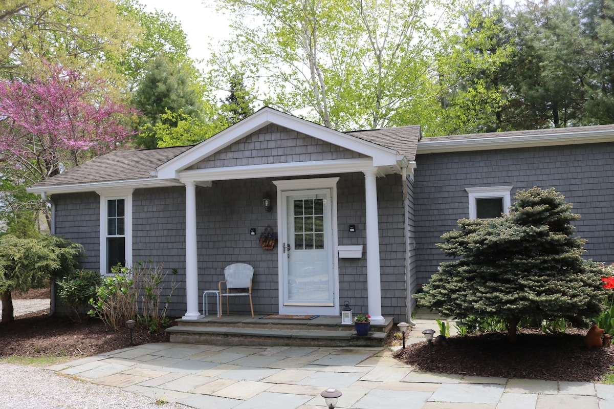 The exterior of a gray cottage is presented, showcasing a covered front porch with a white chair. Stone steps lead to the entryway, framed by greenery and flowering trees. The cottage features a shingle exterior and multiple windows, enhancing the welcoming appearance.