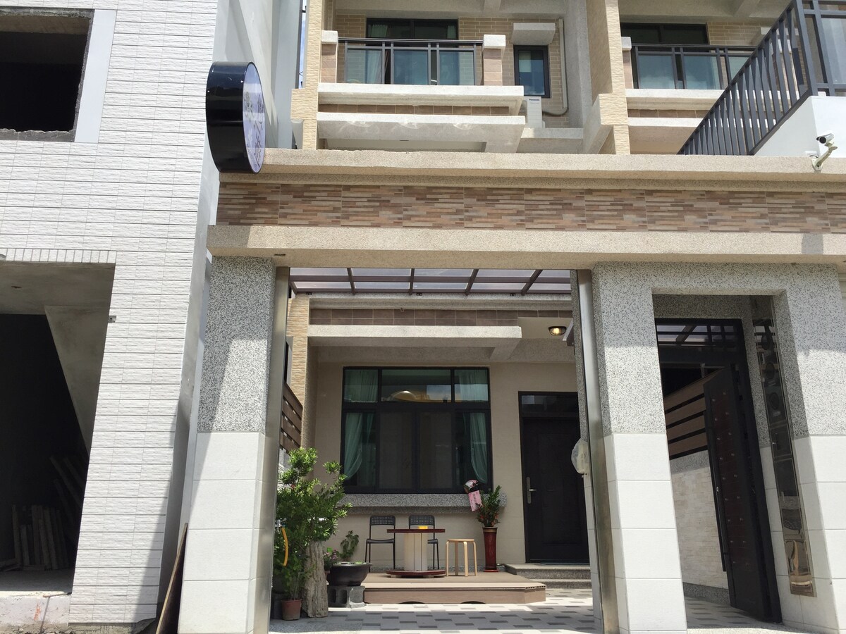 The exterior of the Slowly Inn is displayed, showcasing a modern design with a mix of stone and plaster finishes. The entrance features a covered porch with a small seating area, framed by potted plants. Large windows allow natural light to flow into the interior.