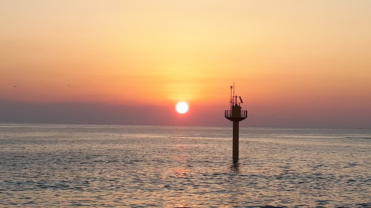 The sun sets on the horizon, casting warm colors across the sky. A solitary lighthouse stands in the water, silhouetted against the vibrant horizon. The calm sea reflects these hues, creating a serene seascape, ideal for relaxation or contemplation.