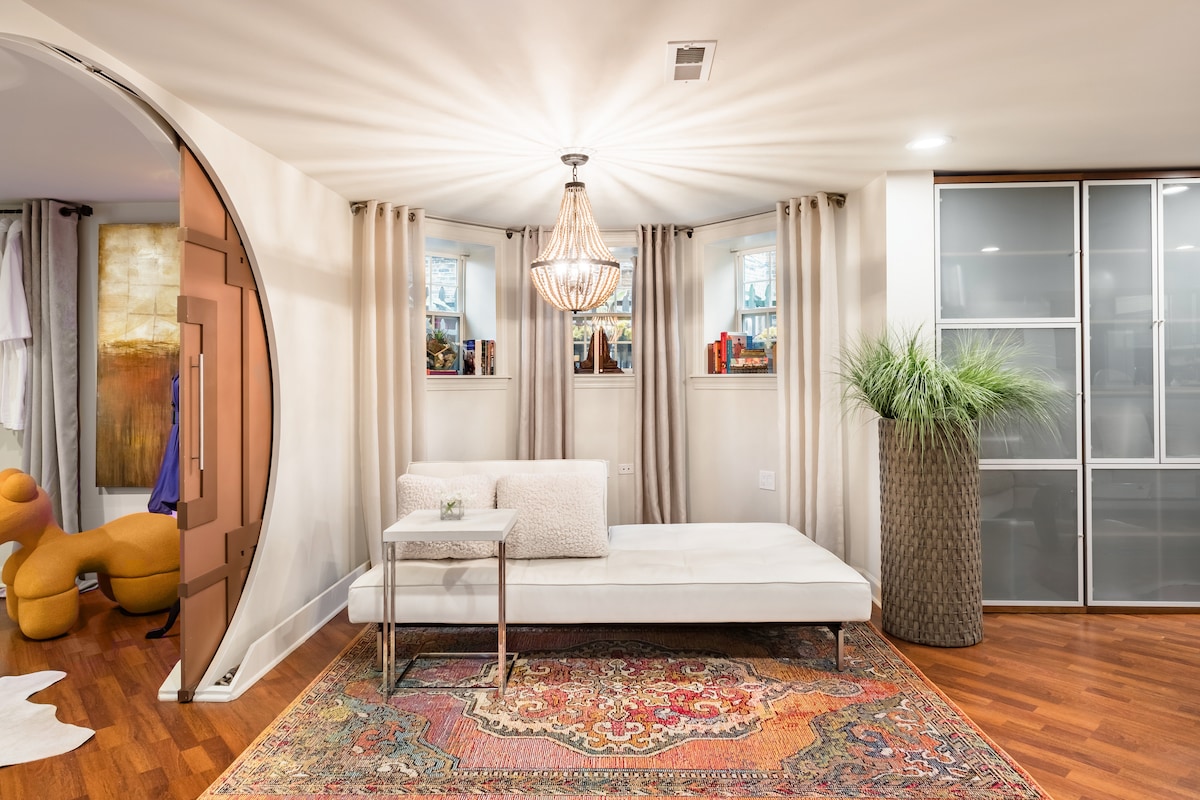 A bright living area features a stylish white sofa positioned on a colorful Persian rug. Elegant lighting is provided by a chandelier, while natural light filters through the windows framed by soft drapes. Decorative plants add a touch of greenery to the space.