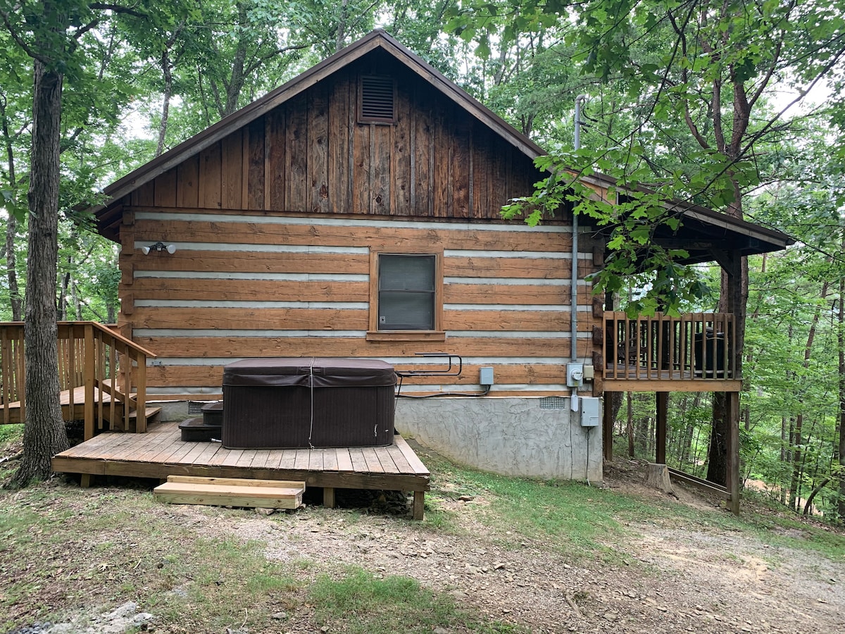 A log cabin exterior is showcased, featuring a wooden deck and a hot tub positioned on the side. Natural surroundings encompass the cabin, with trees providing a sense of privacy. The structure displays classic log construction with horizontal wood accents and a single window.