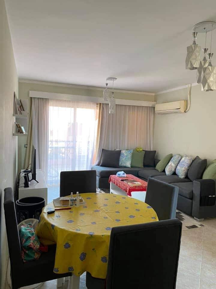 A comfortable living area is showcased, featuring a modern sectional sofa adorned with decorative pillows. A circular dining table with a yellow tablecloth is paired with four gray chairs. Natural light filters in through large glass doors with sheer curtains, enhancing the inviting ambiance.